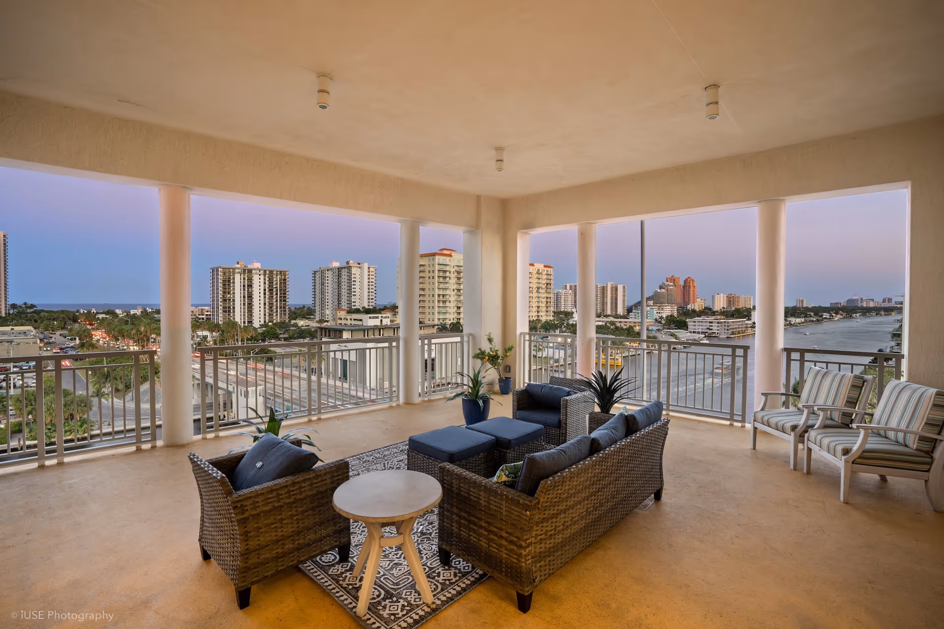A spacious covered outdoor balcony with wicker seating furniture including a sofa, armchair, ottomans, and a small round table on a patterned rug. There are also three striped cushioned chairs along the railing. The balcony overlooks a cityscape with tall buildings and a waterway under a clear sky at dusk.
