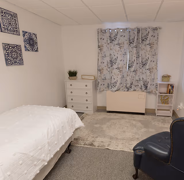 A cozy senior living bedroom with a white bedspread on a single bed, a white chest of drawers with a small plant and framed picture on top, floral curtains covering the window, a light-colored area rug, a small bookshelf with decorative items, and a dark blue upholstered armchair.