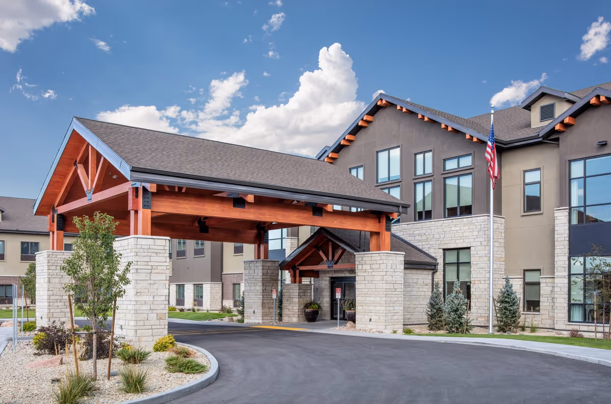 Front entrance of Copper Creek Assisted Living & Memory Care showing a covered porte-cochere with stone pillars, timber beams, and an American flag.