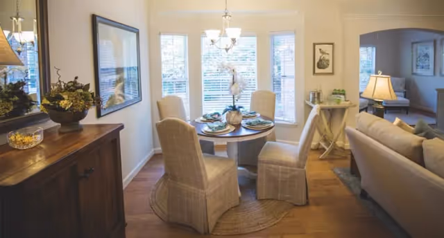 A cozy dining area with a round wooden table set for four, surrounded by four cushioned chairs with plaid slipcovers. The room has large windows with white blinds letting in natural light, a chandelier hanging above the table, and a wooden sideboard with decorative items on the left. Adjacent to the dining area is a living room with a beige sofa, a lamp, and framed artwork on the walls.