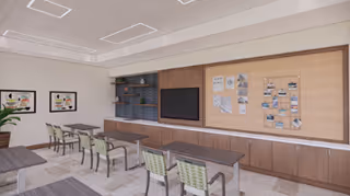 A classroom or meeting room with several tables and green chairs arranged in rows facing a large flat-screen TV mounted on a wooden wall unit. The wall unit also features shelves with decorative items and a large cork bulletin board with various papers and photos pinned on it. The room has a neutral color palette with beige walls, carpeted floor, and ceiling lights.