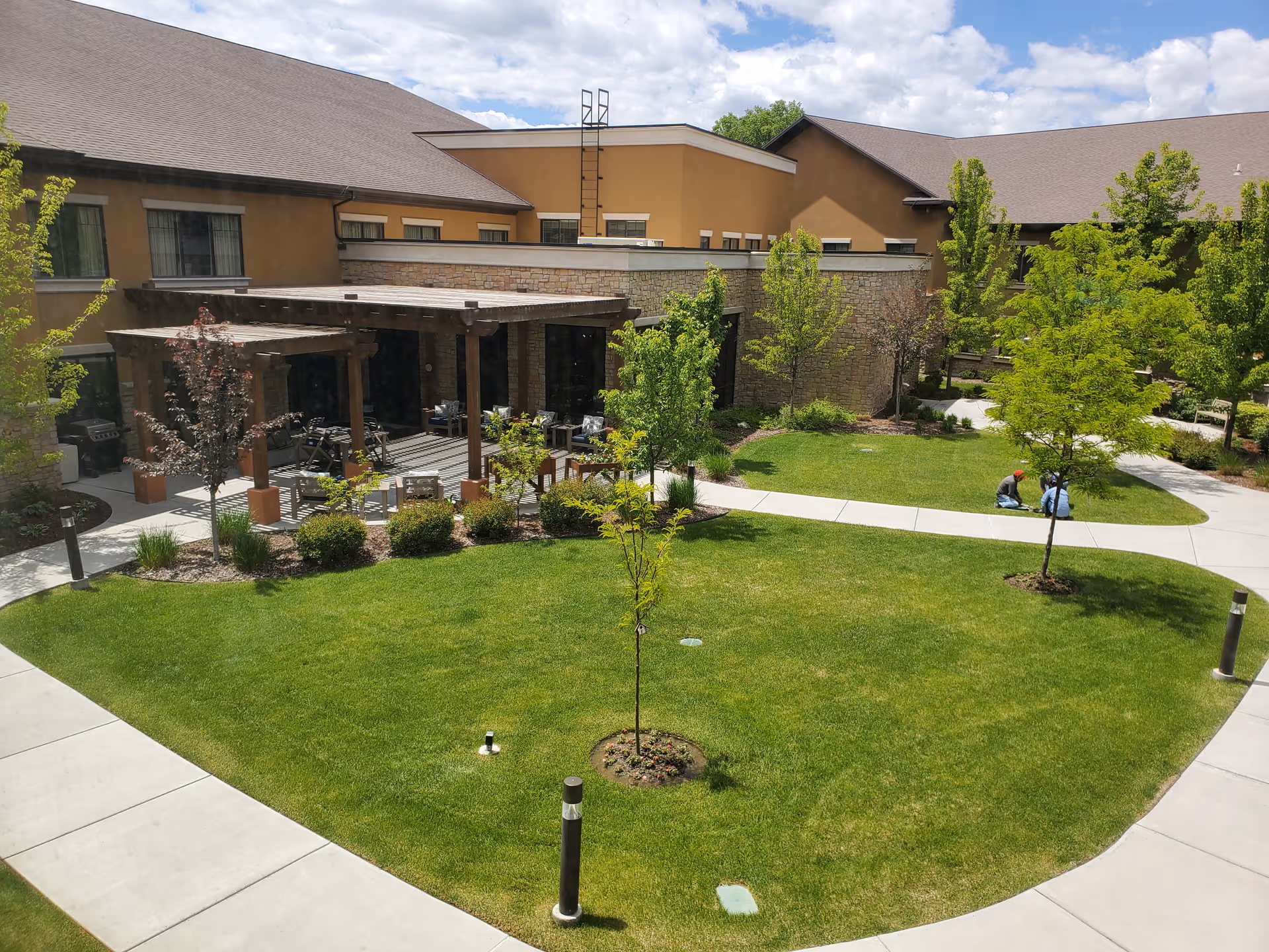 Outdoor courtyard area of The Ridge Cottonwood facility with green grass, young trees, paved walkways, and a shaded seating area under wooden pergolas. Two people are sitting on the grass near the walkway.