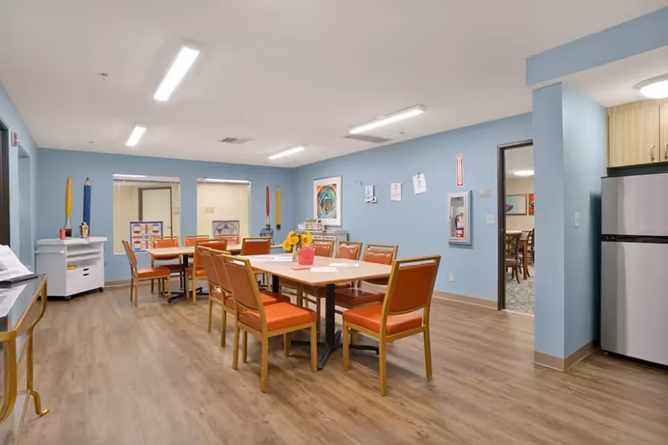 A bright communal dining room with multiple tables and orange chairs in a senior living facility.