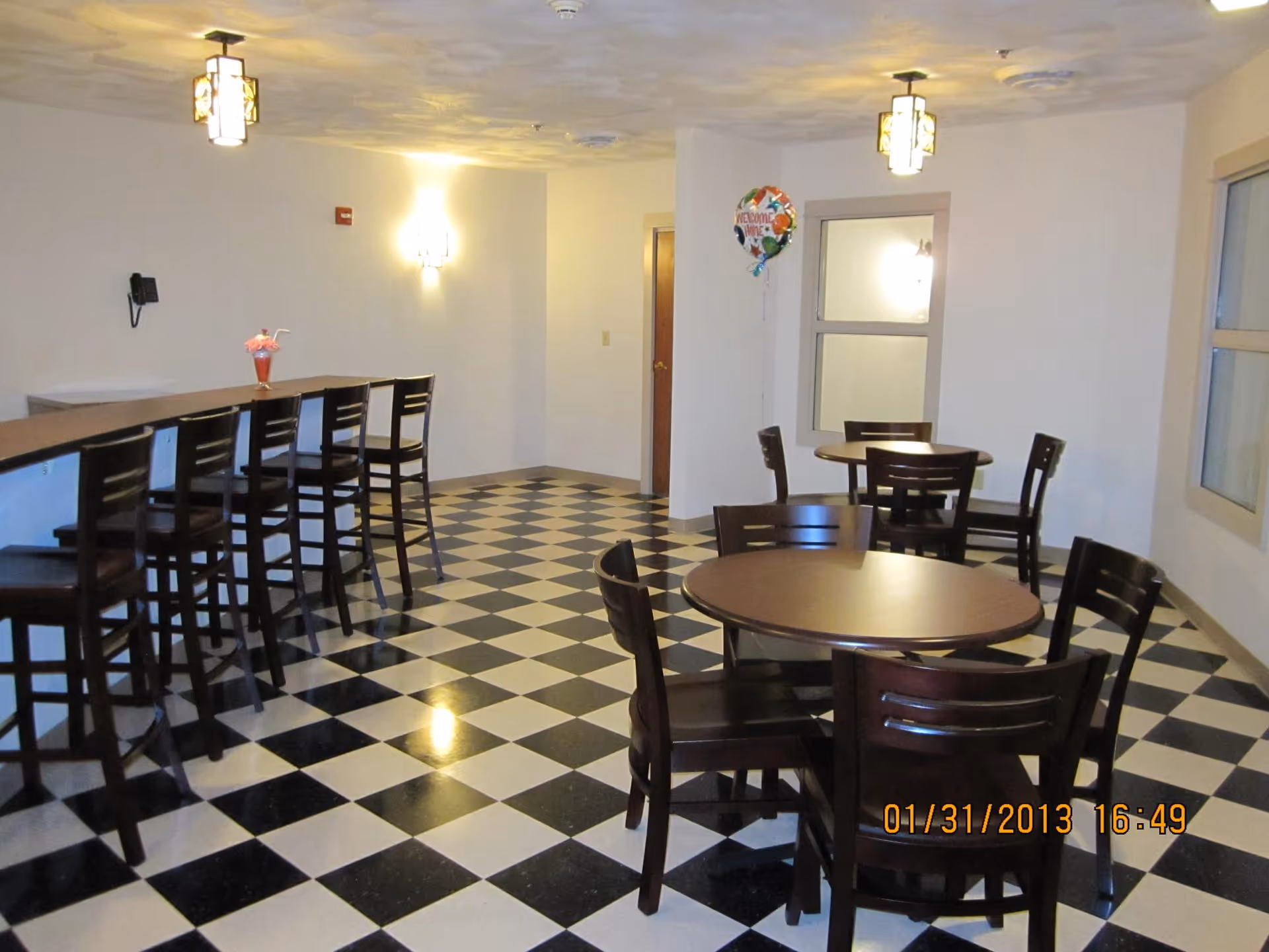 Interior view of a dining area with black and white checkered floor tiles, several round tables with dark wooden chairs, and a counter with high chairs along one wall. The room is lit by ceiling lights and wall sconces, and there is a 'Welcome Home' balloon near a window.