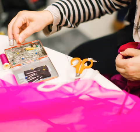 A person wearing a striped sweater is sewing with a needle and thread on a bright pink fabric. Nearby on the table are sewing supplies including a spool of pink thread, a pincushion with many pins, a pair of orange scissors, and a small container of needles.