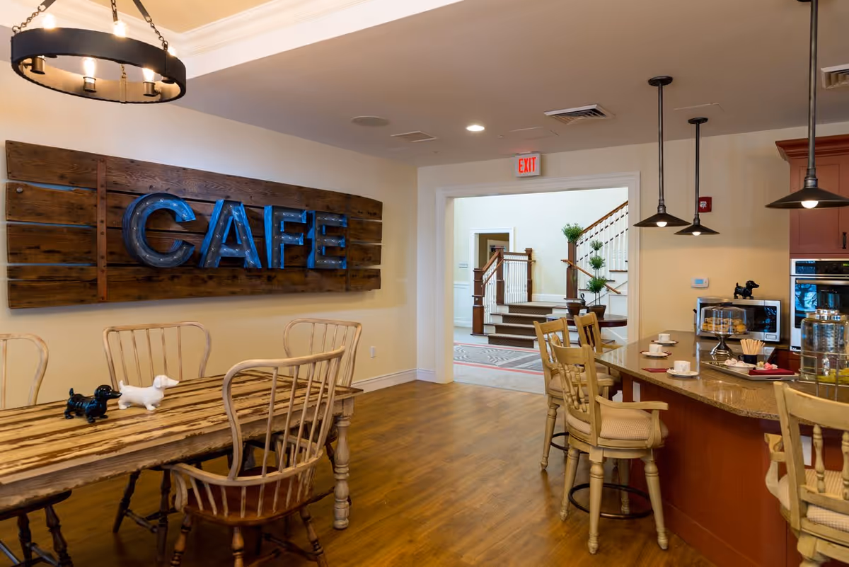 Bright café-style dining area with wooden tables, bar seating and a large blue 'CAFE' sign mounted on a wooden plank wall.