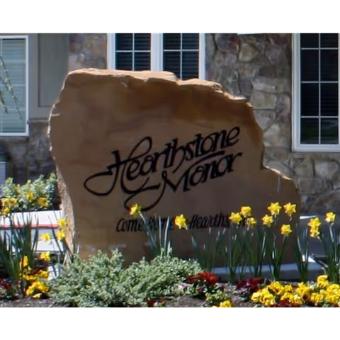 Stone entrance sign reading "Hearthstone Manor" surrounded by daffodils and landscaping in front of a building.