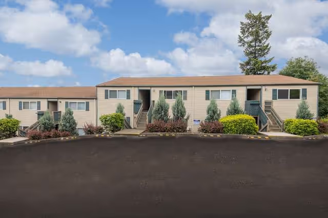Exterior view of a beige, two-story senior living facility with shrubs and a paved parking lot.