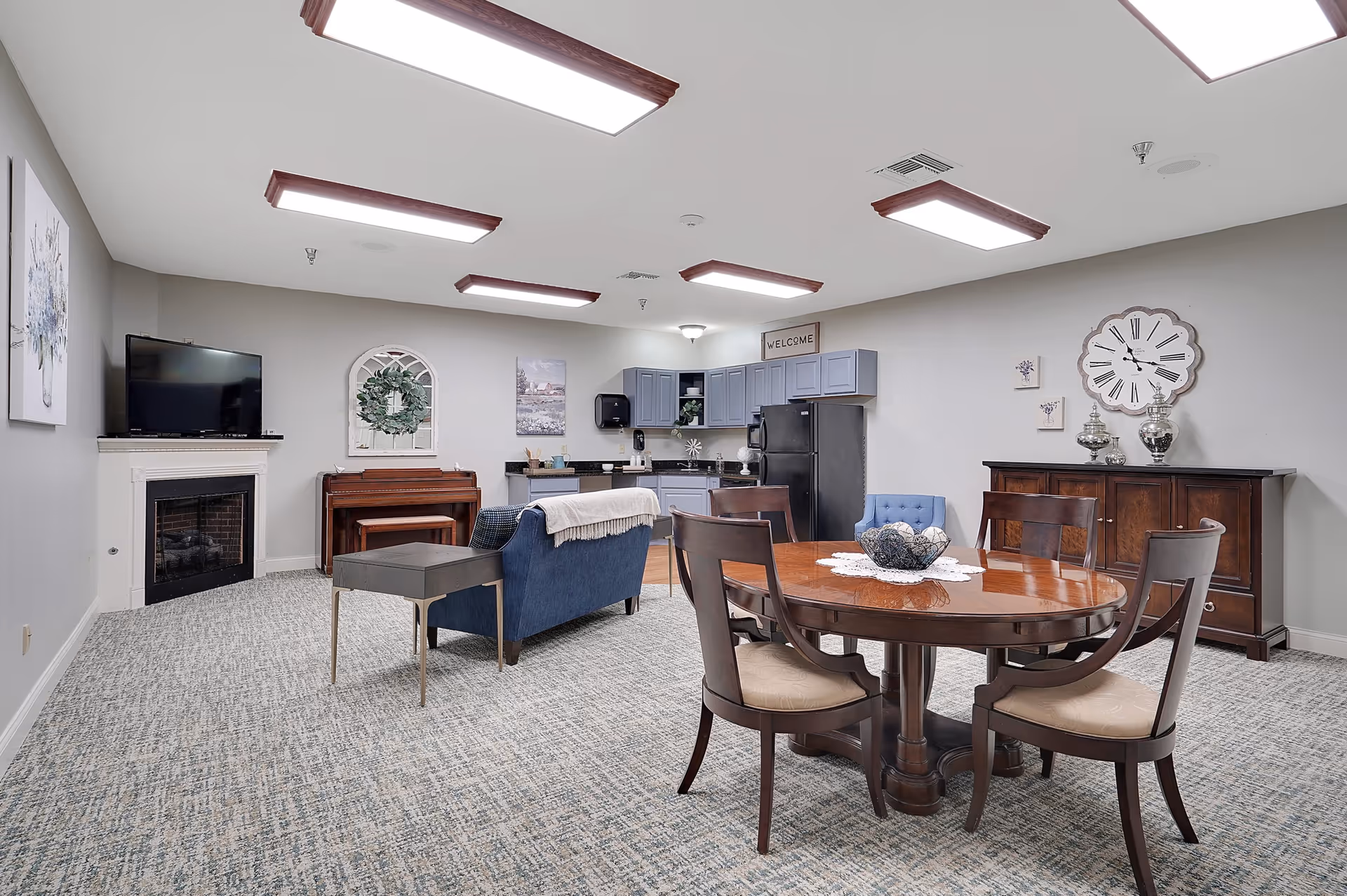 A spacious common area with a round wooden dining table and four chairs in the foreground. Behind the table is a wooden sideboard with decorative items and a large wall clock. To the left, there is a blue couch with a small side table, a piano with a bench, and a fireplace with a TV mounted above it. The back wall features a small kitchenette with gray cabinets, a black refrigerator, and a 'Welcome' sign. The room has carpeted flooring and bright ceiling lights.