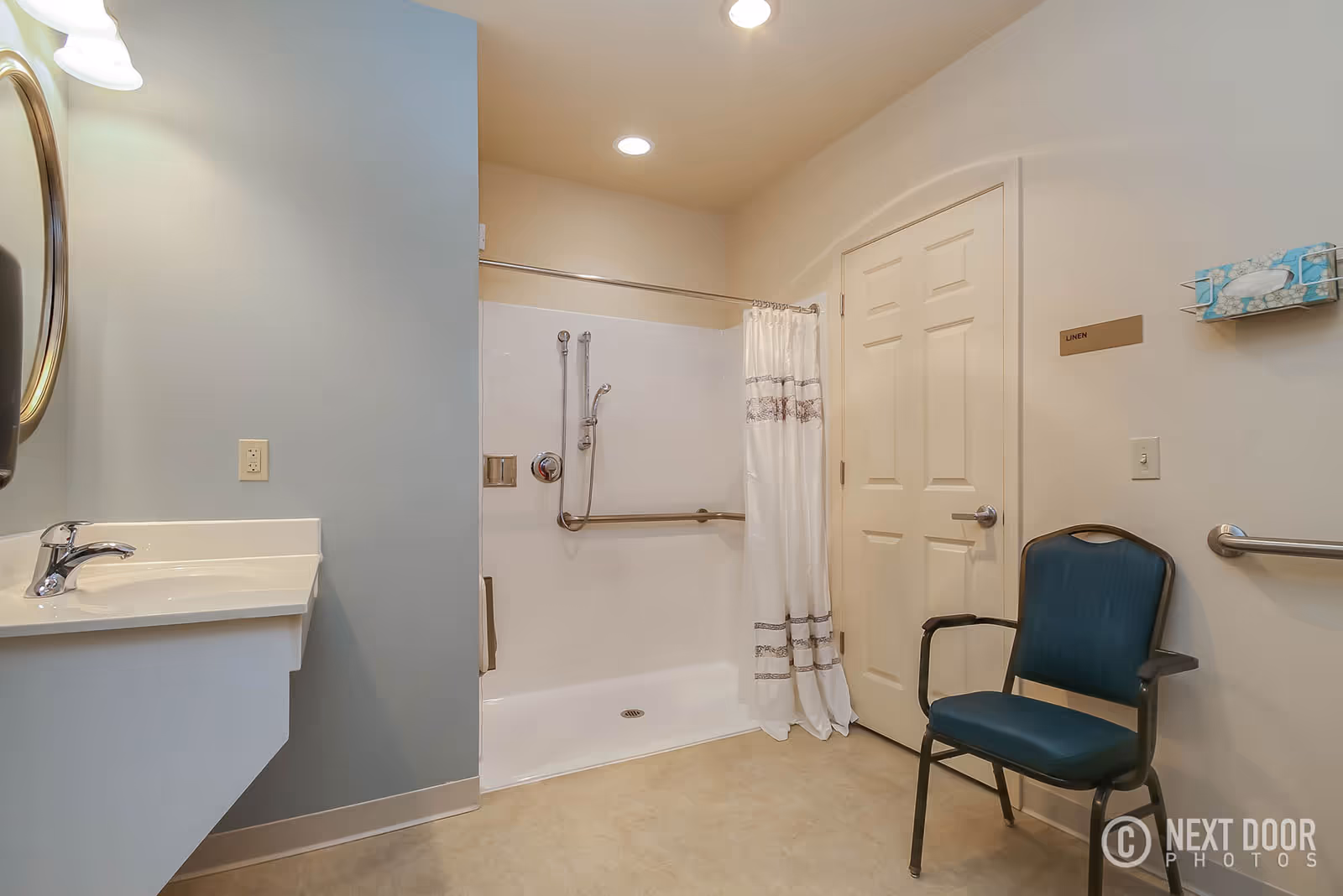 A clean and accessible bathroom with a walk-in shower featuring grab bars and a handheld showerhead. There is a white sink with a chrome faucet and an oval mirror above it. A blue chair with armrests is placed near a door labeled 'Linen'. A tissue box is mounted on the wall above a grab bar.