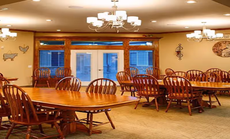 A spacious dining room with multiple wooden tables and matching wooden chairs arranged neatly. The room features beige walls, a large clock on the wall, decorative wall hangings, and multiple chandeliers providing warm lighting. There are large windows and a glass door at the far end of the room.