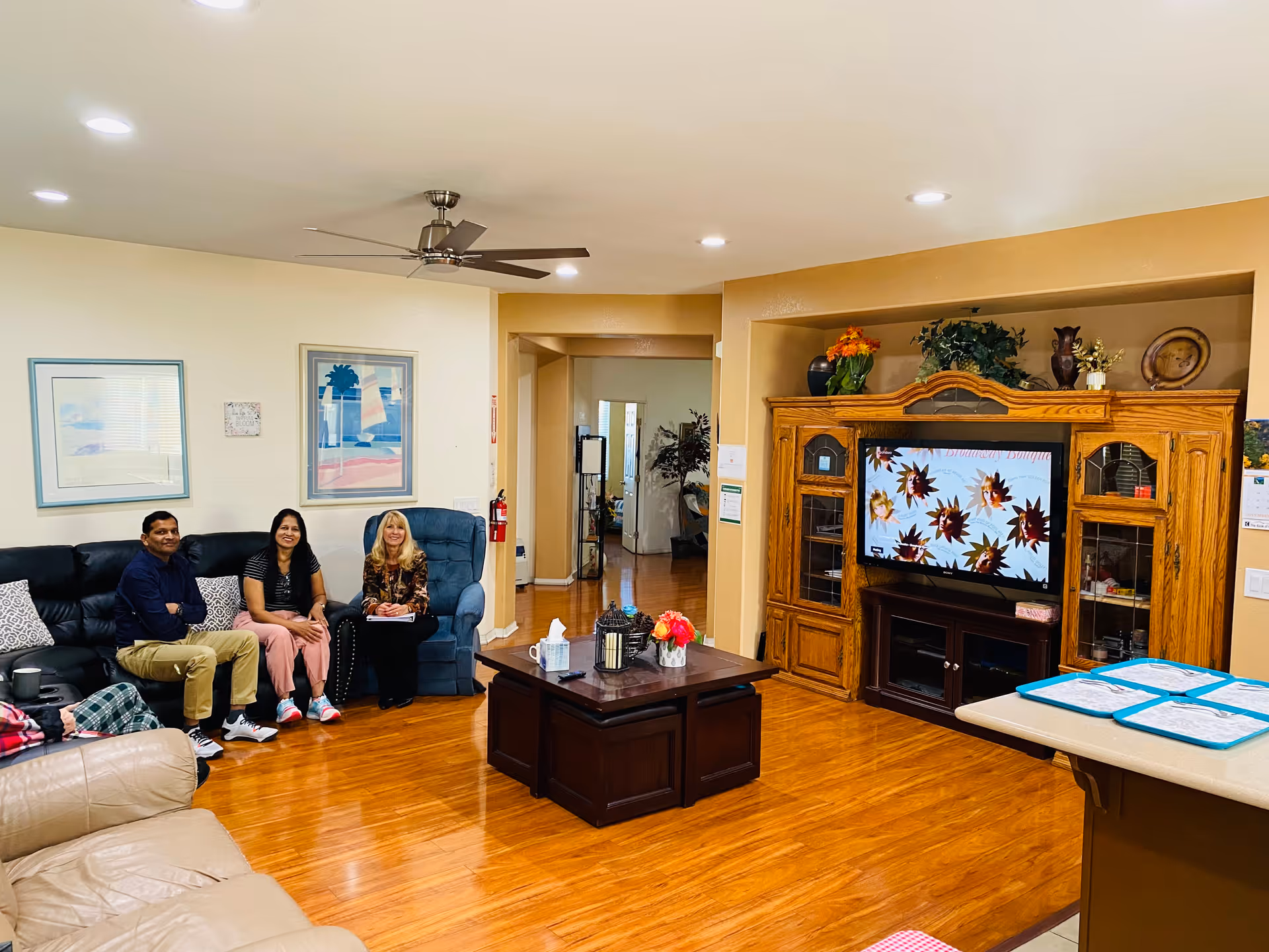 A cozy living room with three people sitting on a black leather couch and a blue armchair. The room has wooden flooring, a ceiling fan, and recessed lighting. There is a wooden entertainment center with a TV displaying a floral pattern. A coffee table with decorative items is in the center, and the walls are adorned with framed artwork.