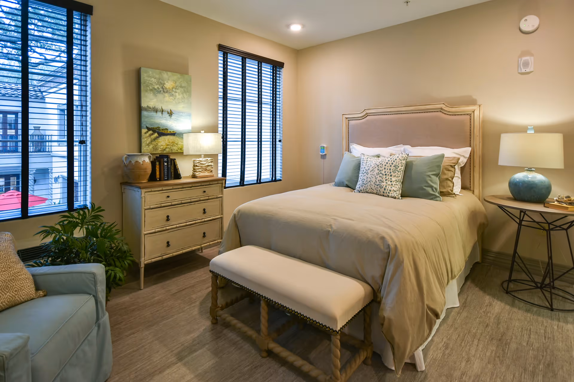 A cozy bedroom with a beige upholstered bed adorned with multiple pillows, a cushioned bench at the foot of the bed, a wooden dresser with books and a lamp, a round side table with a blue lamp, a light blue armchair, and two large windows with black blinds letting in natural light.
