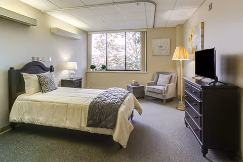 A well-lit bedroom in a senior living facility featuring a single bed with white and gray bedding, a nightstand with a lamp, a cushioned armchair next to a small round table, a floor lamp, a dresser with a flat-screen TV, and a large window with a view of trees outside.