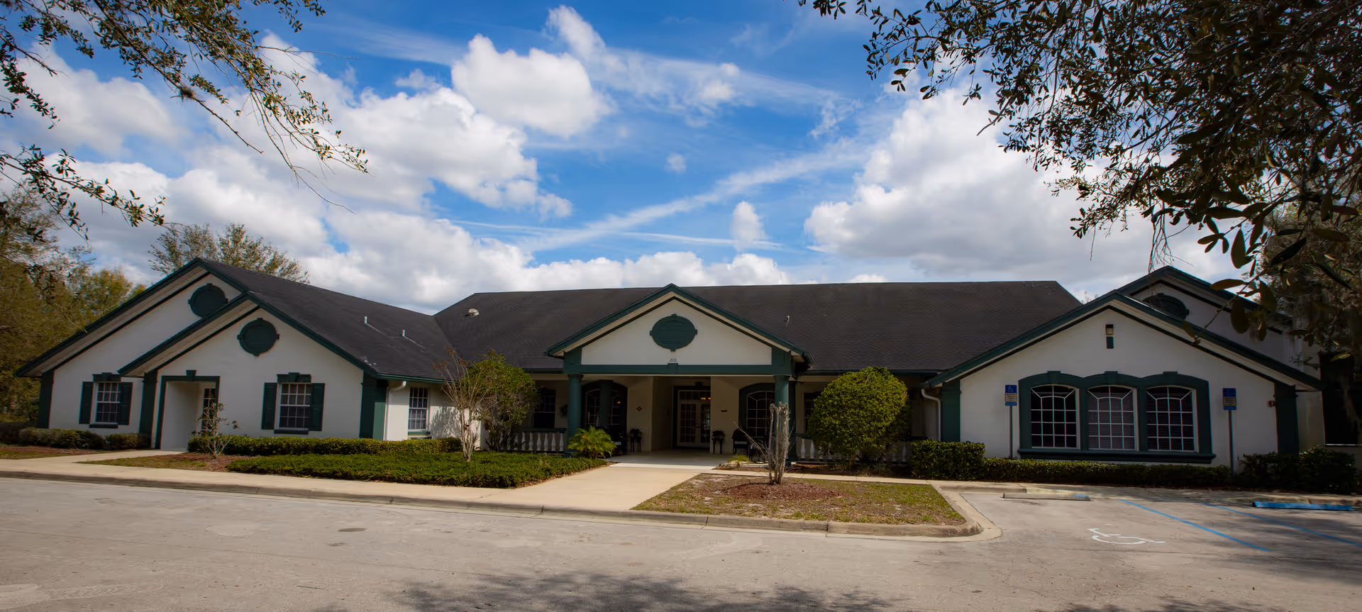 Front exterior view of a single-story building with white walls and green trim, featuring a central entrance with a covered porch, surrounded by bushes and trees under a partly cloudy blue sky.