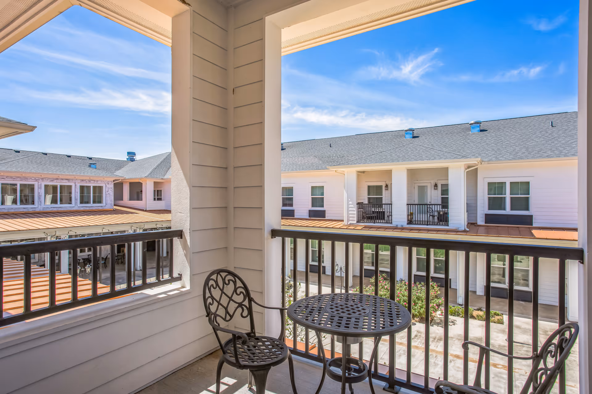 View from a covered balcony with a small round metal table and two matching chairs, overlooking the courtyard of a senior living facility with white buildings, windows, and a clear blue sky.