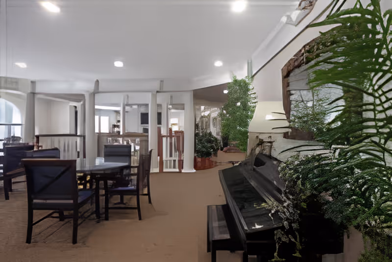 Interior view of a senior living facility common area with several tables and chairs arranged for seating. A black upright piano is positioned on the right side, surrounded by green plants. The space features white walls, columns, and a carpeted floor, with a bright and airy atmosphere.