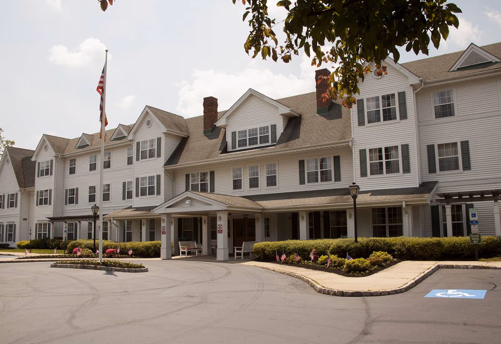 Front exterior of a multi-story white senior living building with a covered entrance, flagpole, landscaped driveway, and handicap parking.