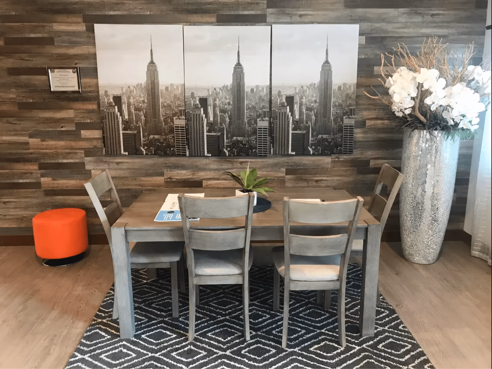 A small dining area with a wooden table and four chairs against a wood-paneled wall featuring a black-and-white city skyline triptych, a patterned rug, a tall vase of white flowers, and an orange ottoman.