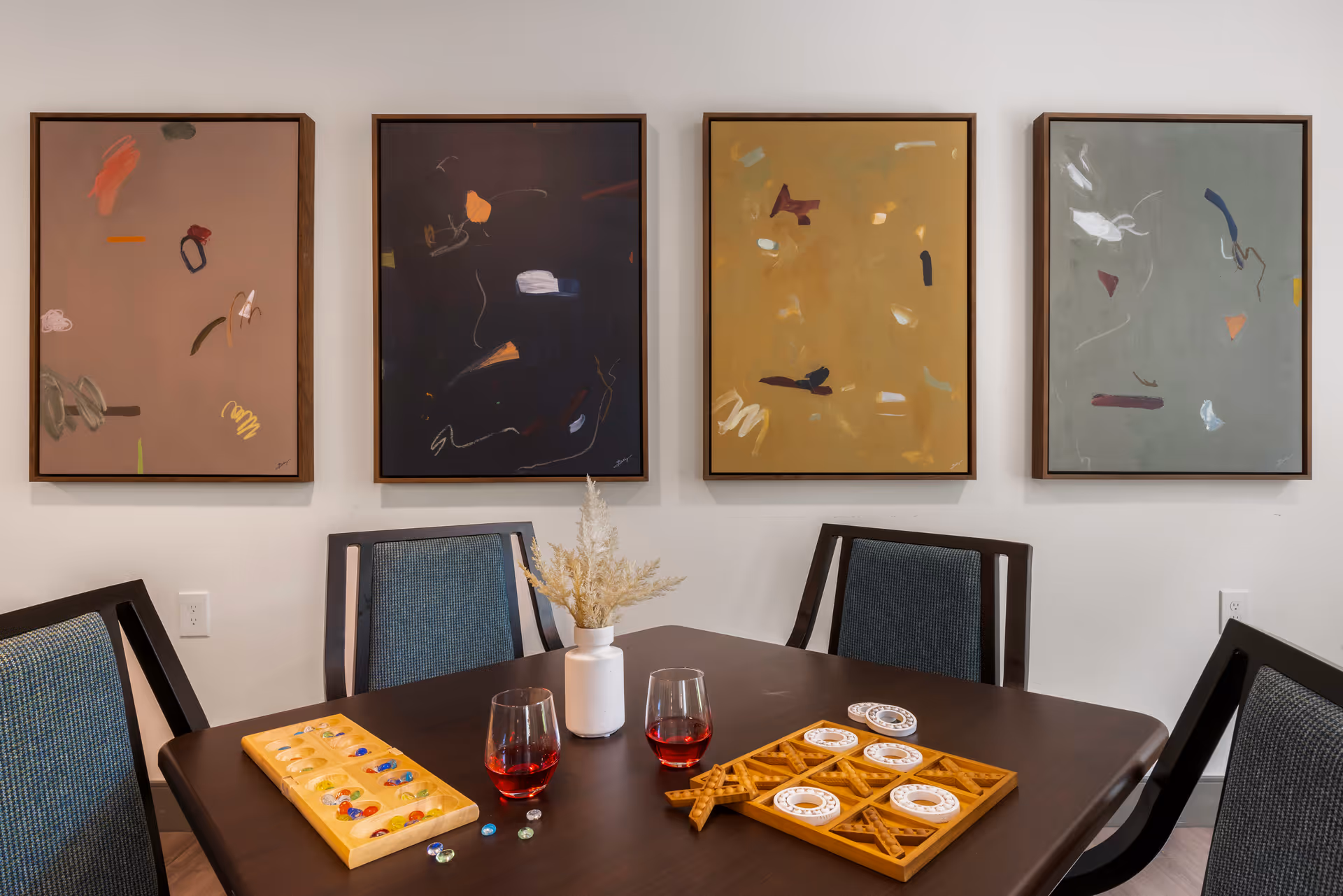 A dining area with a dark wooden table and four chairs with blue upholstery. On the table are two glasses with red liquid, a wooden board game with colorful pieces, a tic-tac-toe game, and a white vase with dried flowers. Four abstract paintings in different colors hang on the white wall behind the table.