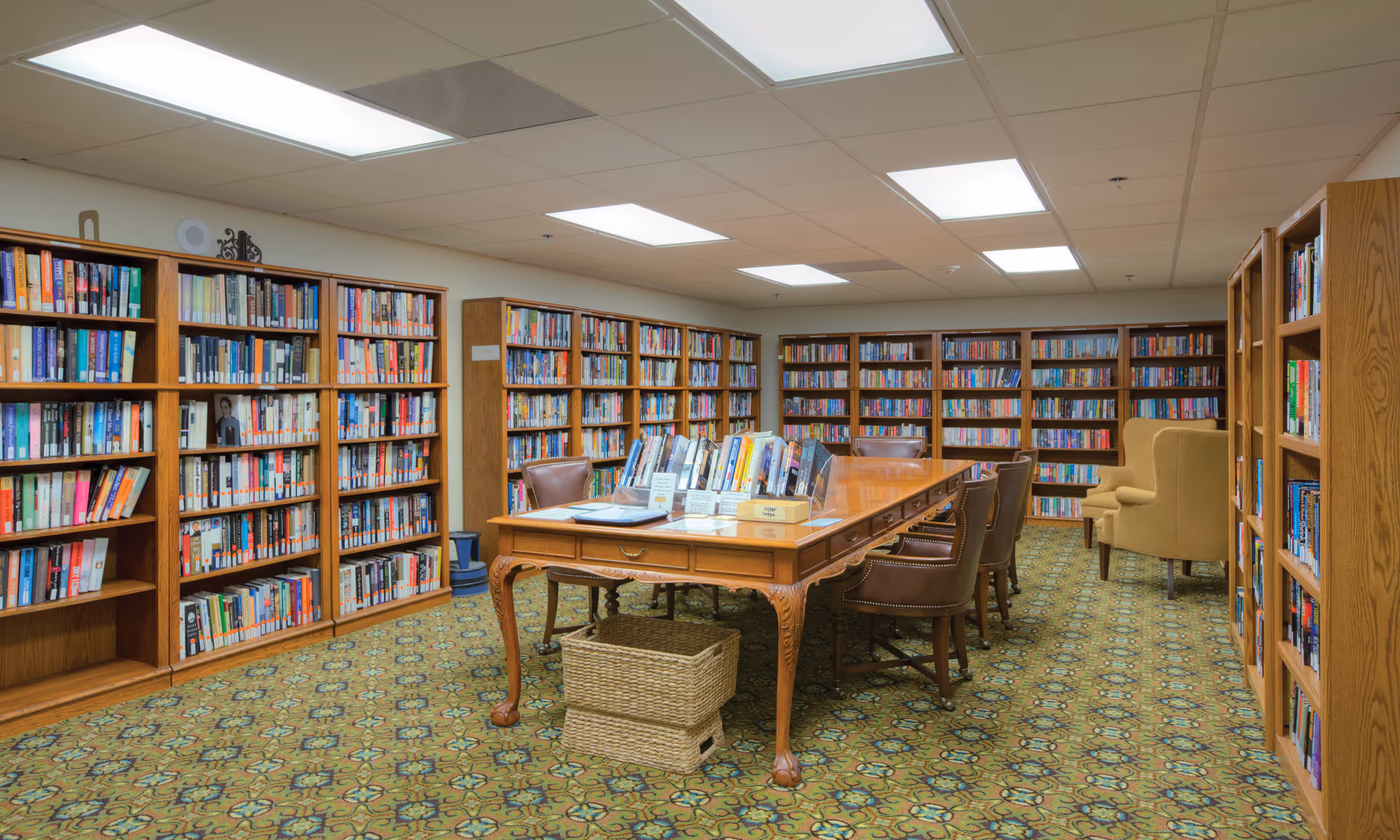 A well-lit library room with wooden bookshelves filled with books lining the walls. In the center, there is a large wooden table with several chairs around it and books displayed on top. Two beige armchairs are positioned near the back corner. The floor is covered with a patterned carpet.