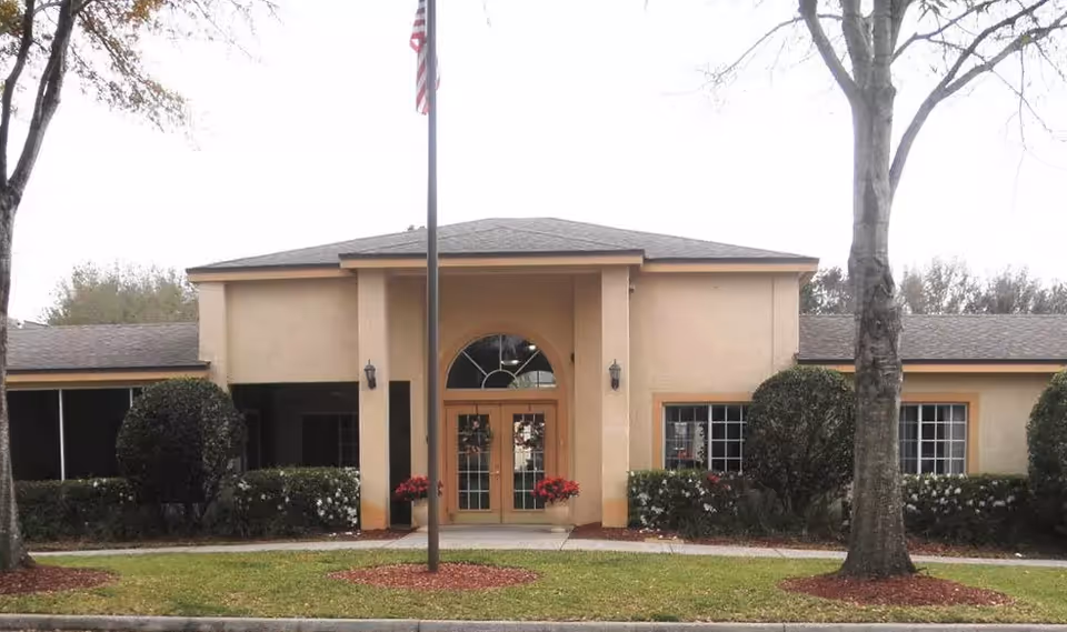 Front entrance of a beige stucco building with double glass doors, a flagpole, and landscaped shrubs and trees.