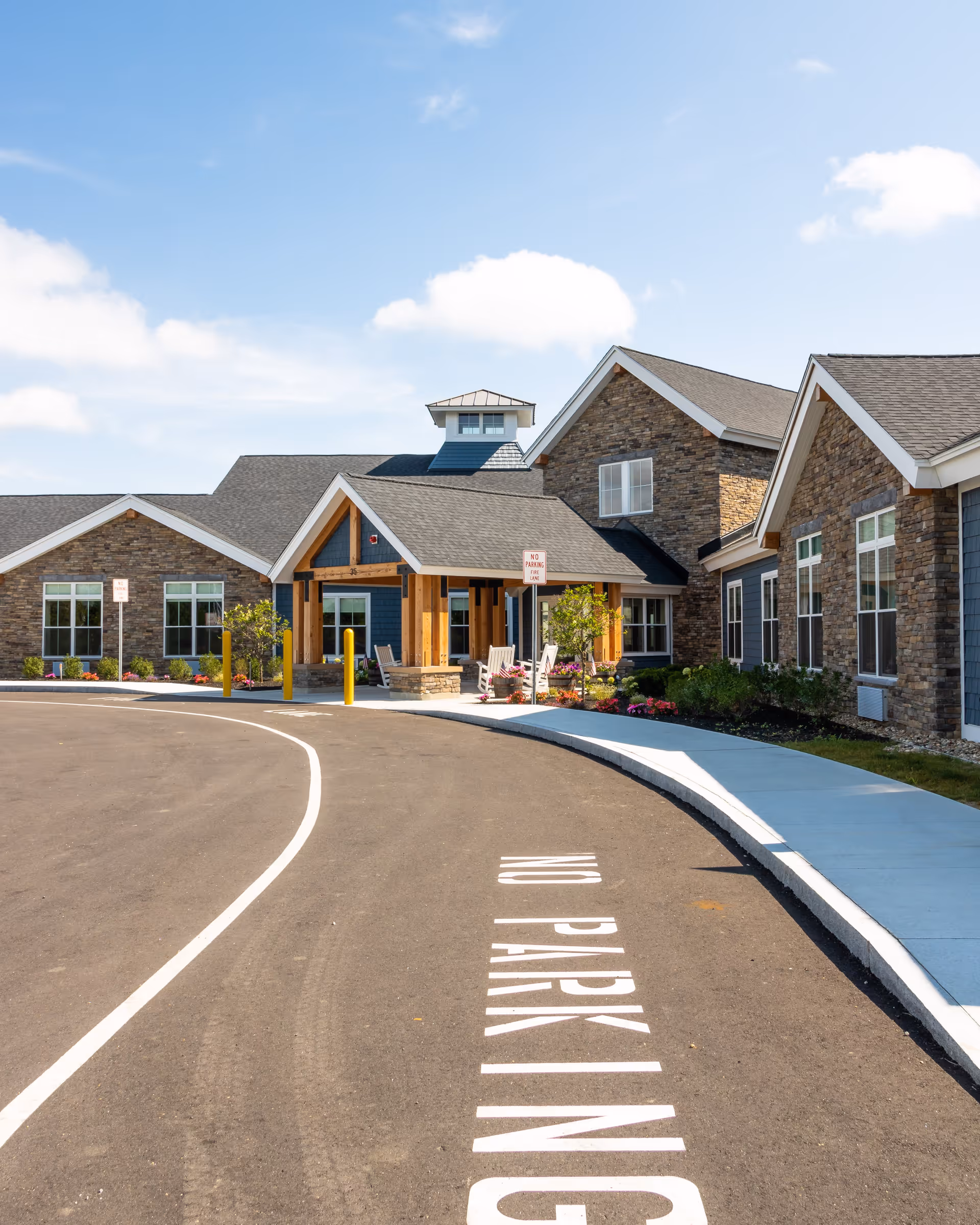 Exterior view of a senior living facility named Spring Village at Dover, showing a curved driveway with 'NO PARKING' painted on the asphalt, a covered entrance with wooden beams, stone and siding exterior walls, several windows, and a clear blue sky with some clouds.