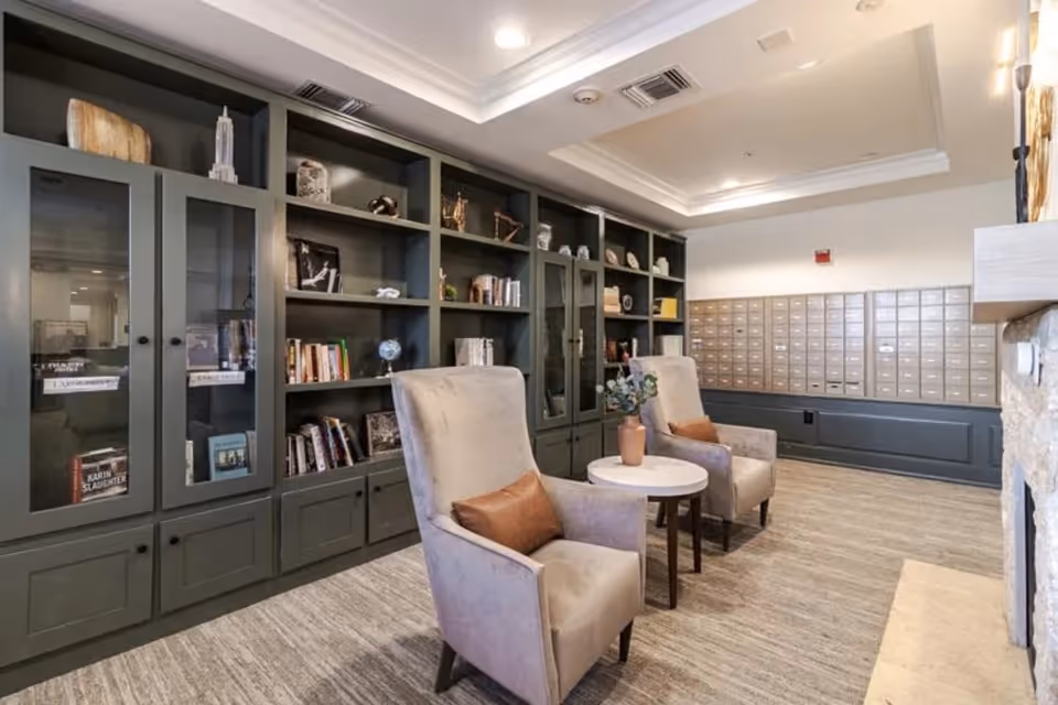 A cozy interior room featuring two beige armchairs with brown pillows positioned around a small round table with a vase of flowers. Behind the chairs is a large built-in bookshelf filled with books and decorative items. To the right, there is a wall of mailboxes. The room has a light-colored carpet and a tray ceiling with recessed lighting.