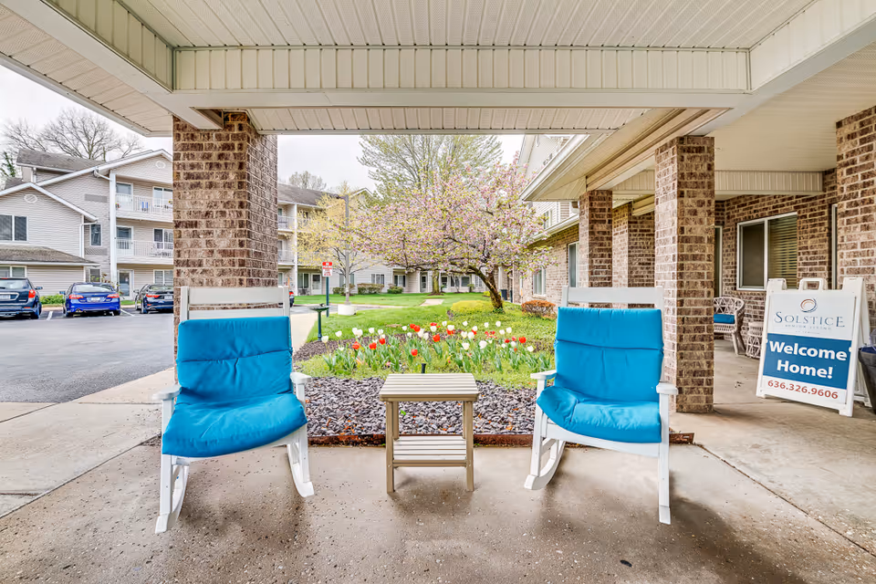 Covered entrance of a senior living building with two blue-cushioned chairs, a small table, and a tulip flowerbed in front of the complex.