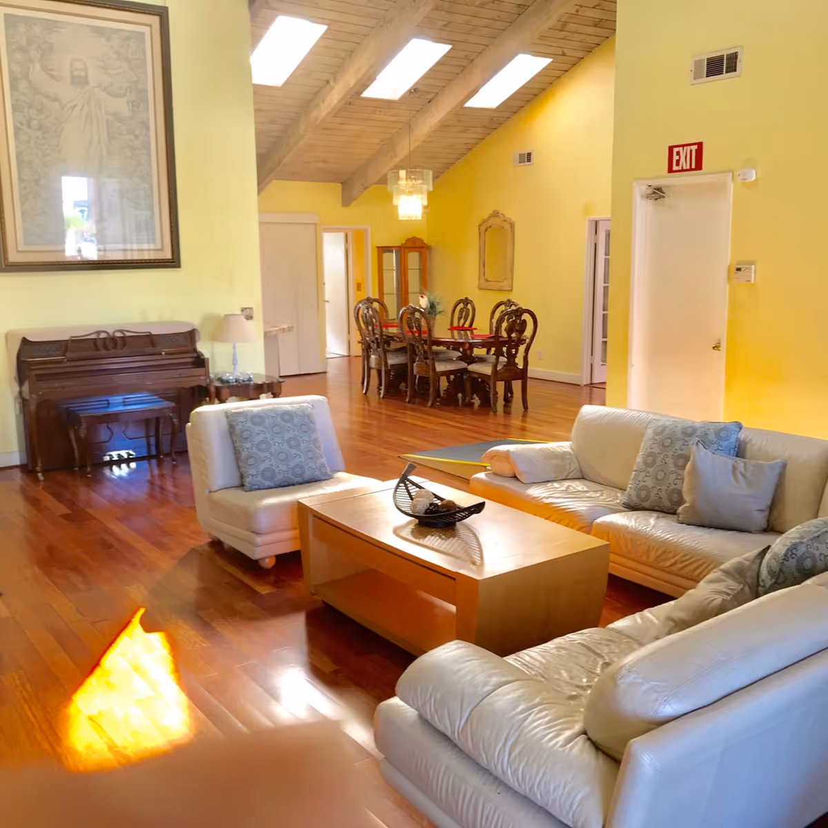 Bright living room with vaulted skylit ceiling, leather sofas and a coffee table in the foreground, a piano to the left and a dining table in the background.