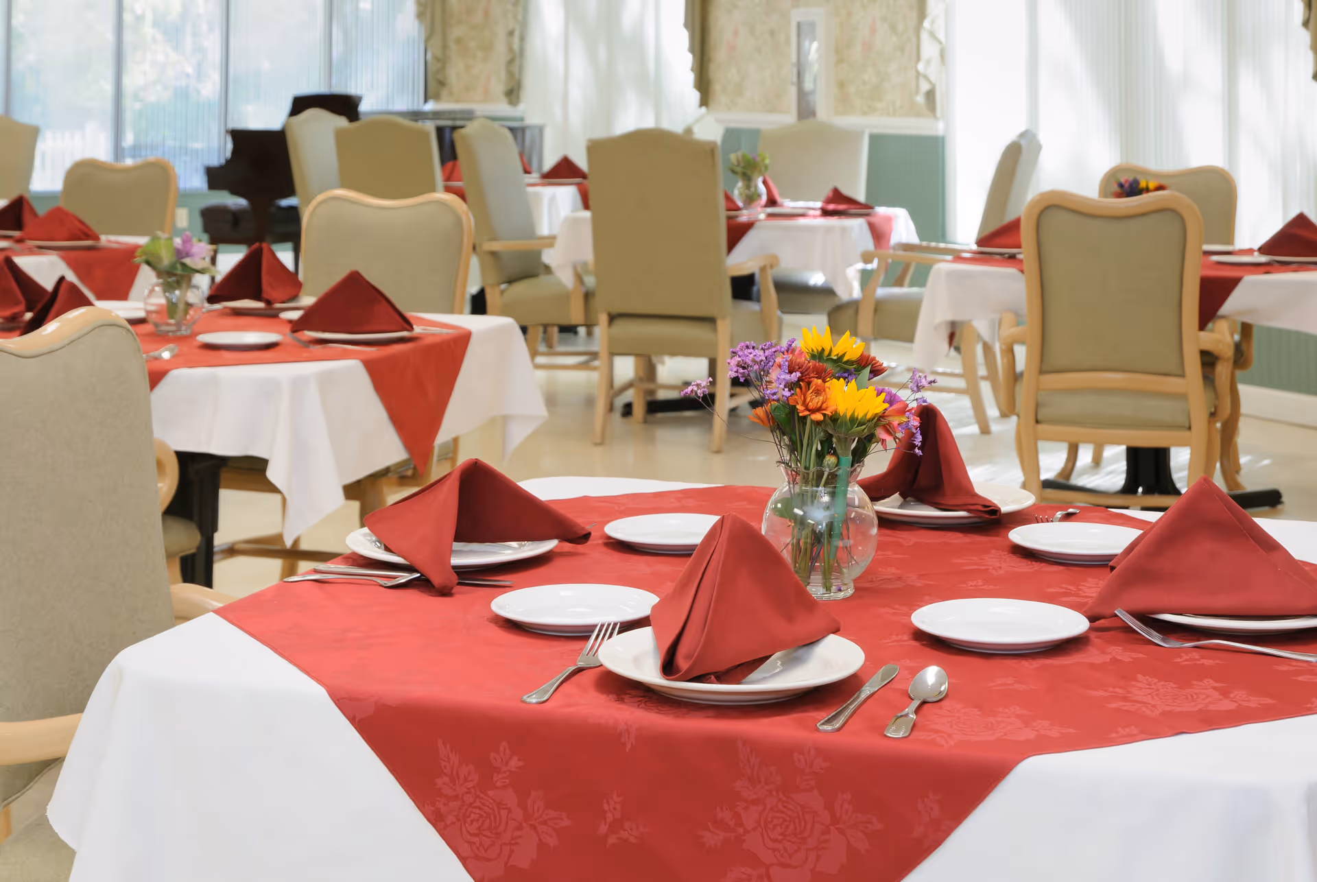 Dining room with round tables set with red napkins, white plates and a vase of flowers, surrounded by upholstered chairs.