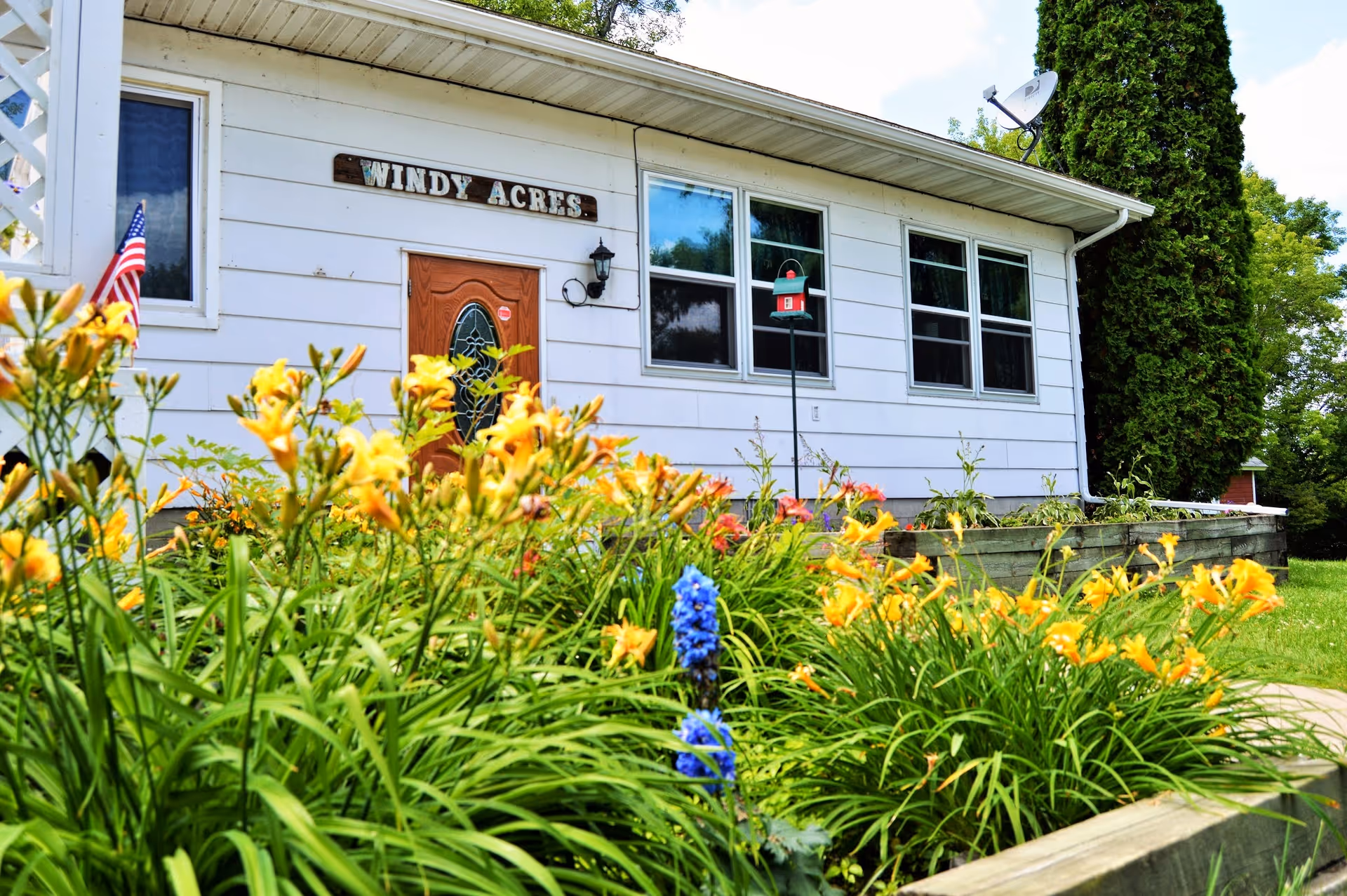 Exterior view of Windy Acres Long Term Care Inc. building with white siding, a wooden door with decorative glass, and three windows. In the foreground, there is a garden with yellow and purple flowers, green plants, and an American flag on the left side.