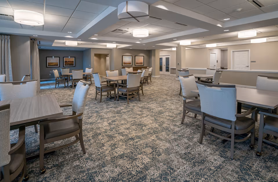 A spacious dining room in a senior living facility with multiple wooden tables and cushioned chairs arranged neatly on a patterned carpet. The room is well-lit with modern ceiling lights and has neutral-colored walls with framed artwork. There are windows with curtains on the left side and doors at the far end of the room.