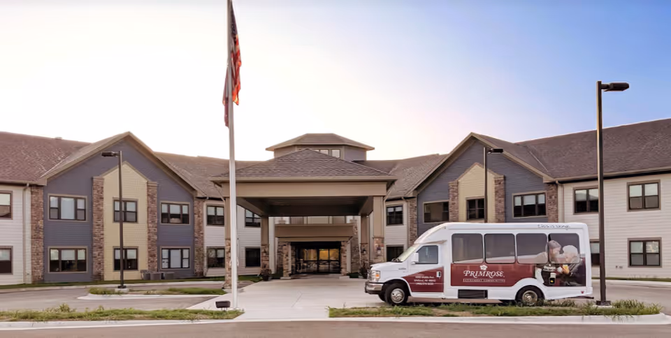 Front exterior view of Primrose Retirement Community of Midland building with a covered entrance, an American flag on a flagpole, and a white shuttle van parked in front displaying the Primrose logo and images of elderly people.