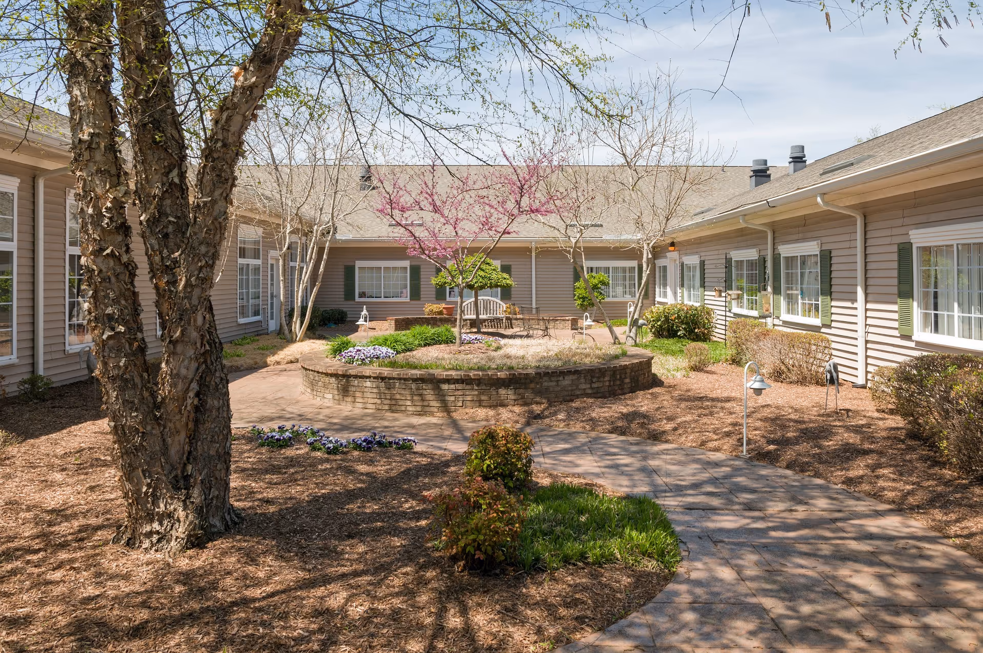 Sunny landscaped courtyard with a circular brick planter, benches, and surrounding single-story facility windows.