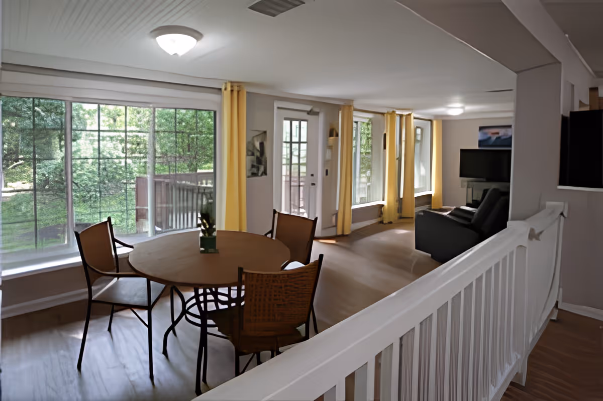 Sunlit open living and dining area with a round table and chairs, a sofa and TV, and large windows with yellow curtains.