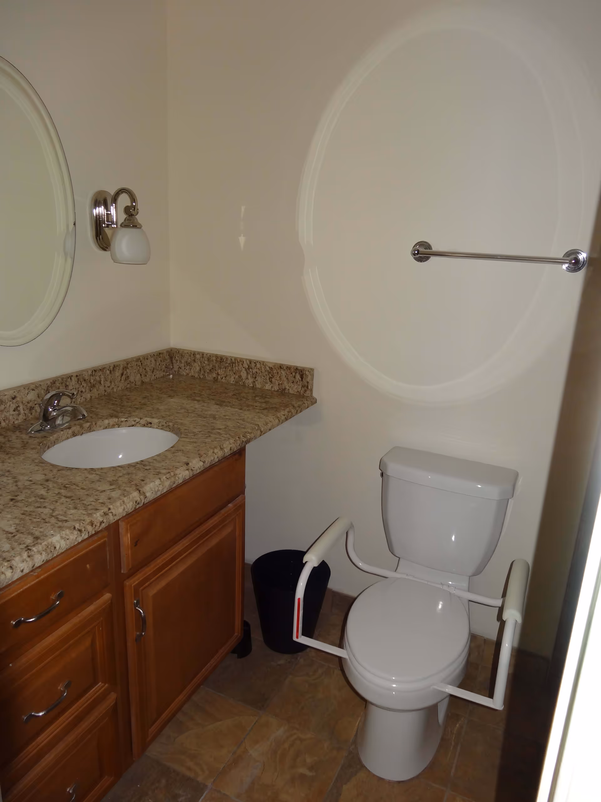 A bathroom with a white toilet equipped with safety rails on both sides, a wooden vanity with a granite countertop and an oval sink, a round mirror above the sink, a wall-mounted light fixture, a towel bar on the wall, and a small black trash bin on the floor.
