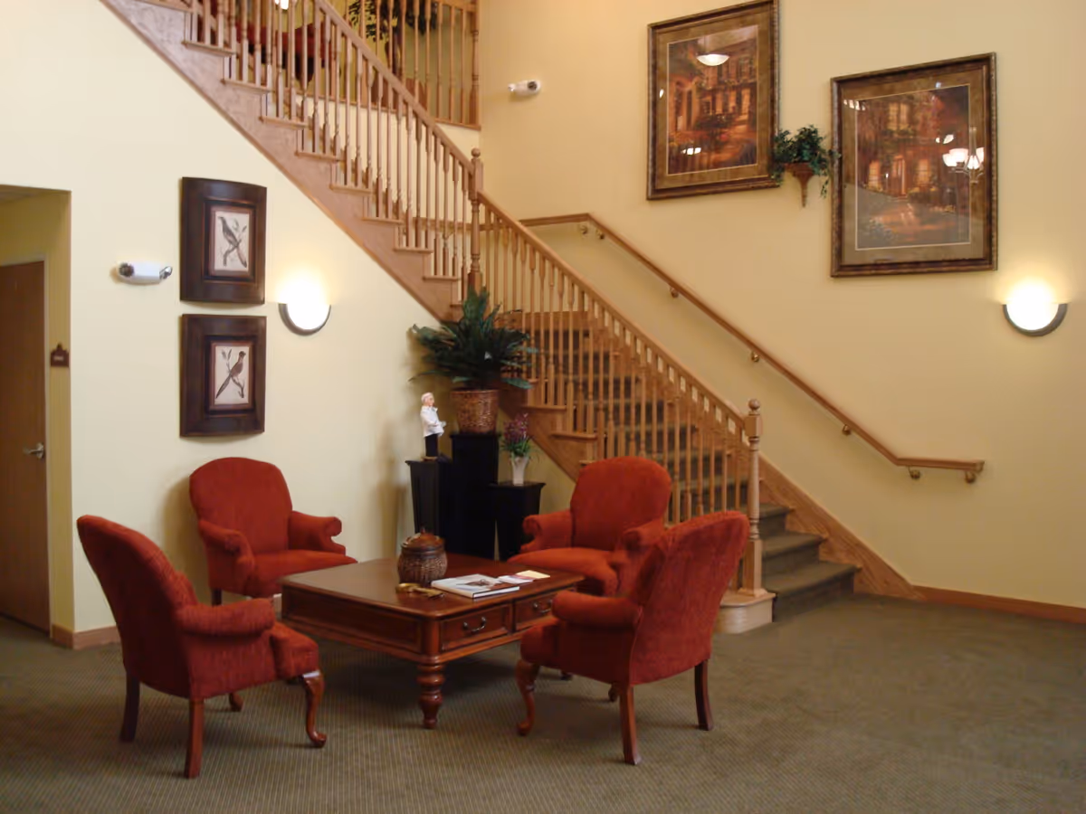 A cozy seating area with four red upholstered armchairs arranged around a wooden coffee table in front of a staircase with wooden railings. The walls are painted light yellow and decorated with framed artwork and wall sconces. There are also potted plants and decorative items near the staircase.