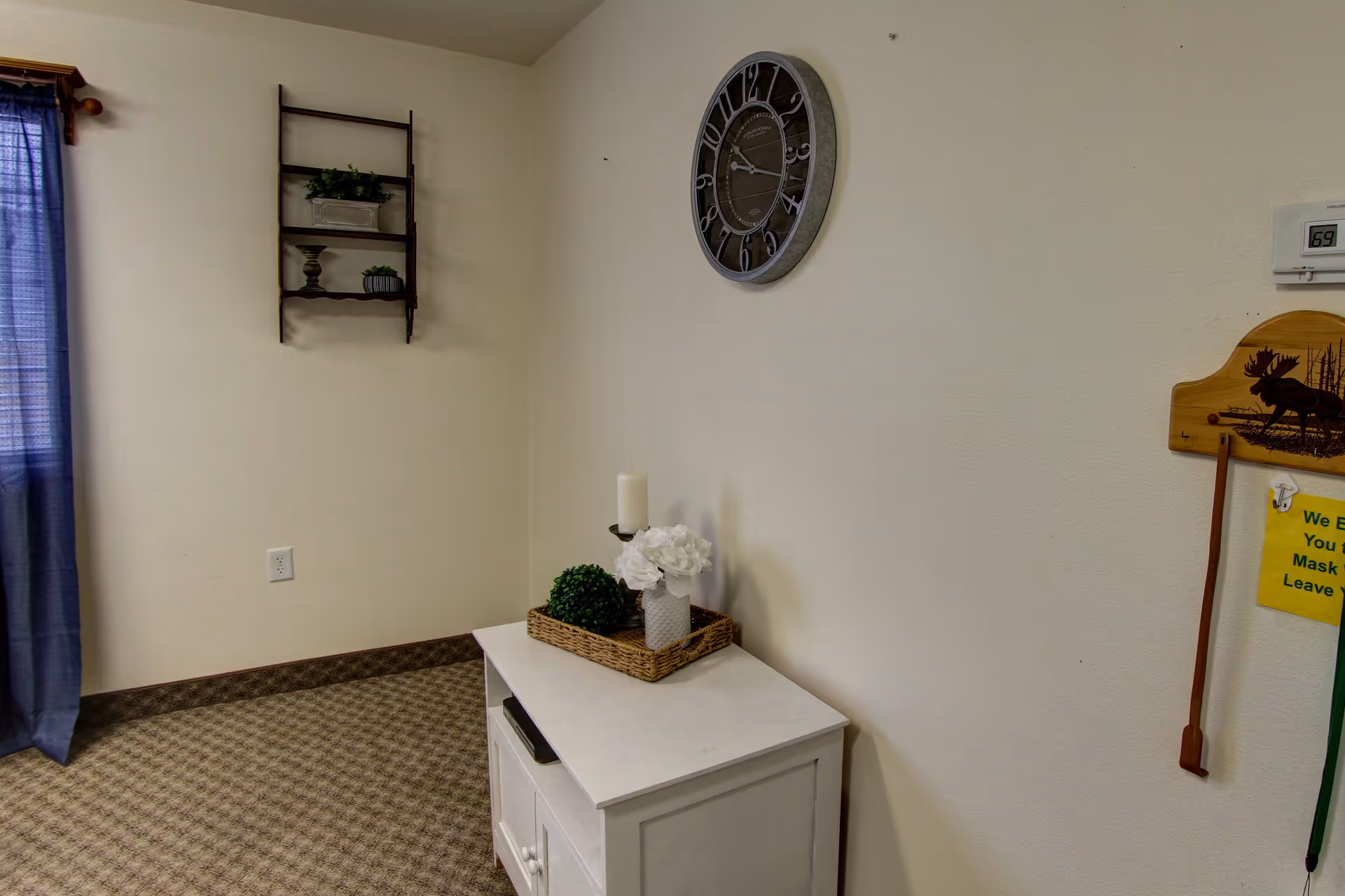 Corner of a decorated room with a small white cabinet topped with a wicker tray and decor, a wall clock, a small wall shelf, and a curtain.