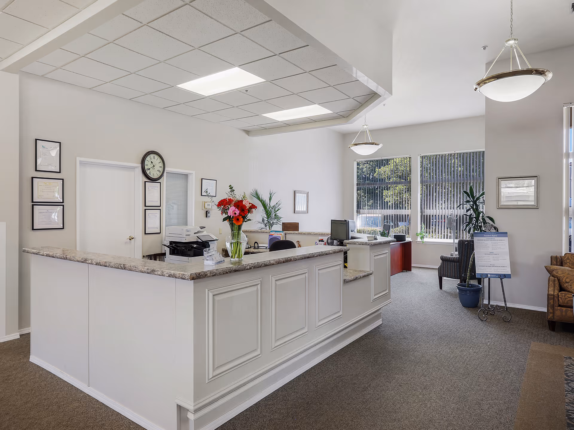 Reception area of a senior living facility with a white front desk, a vase of colorful flowers on the counter, office equipment, framed certificates on the wall, and large windows letting in natural light. There are chairs and plants in the waiting area.