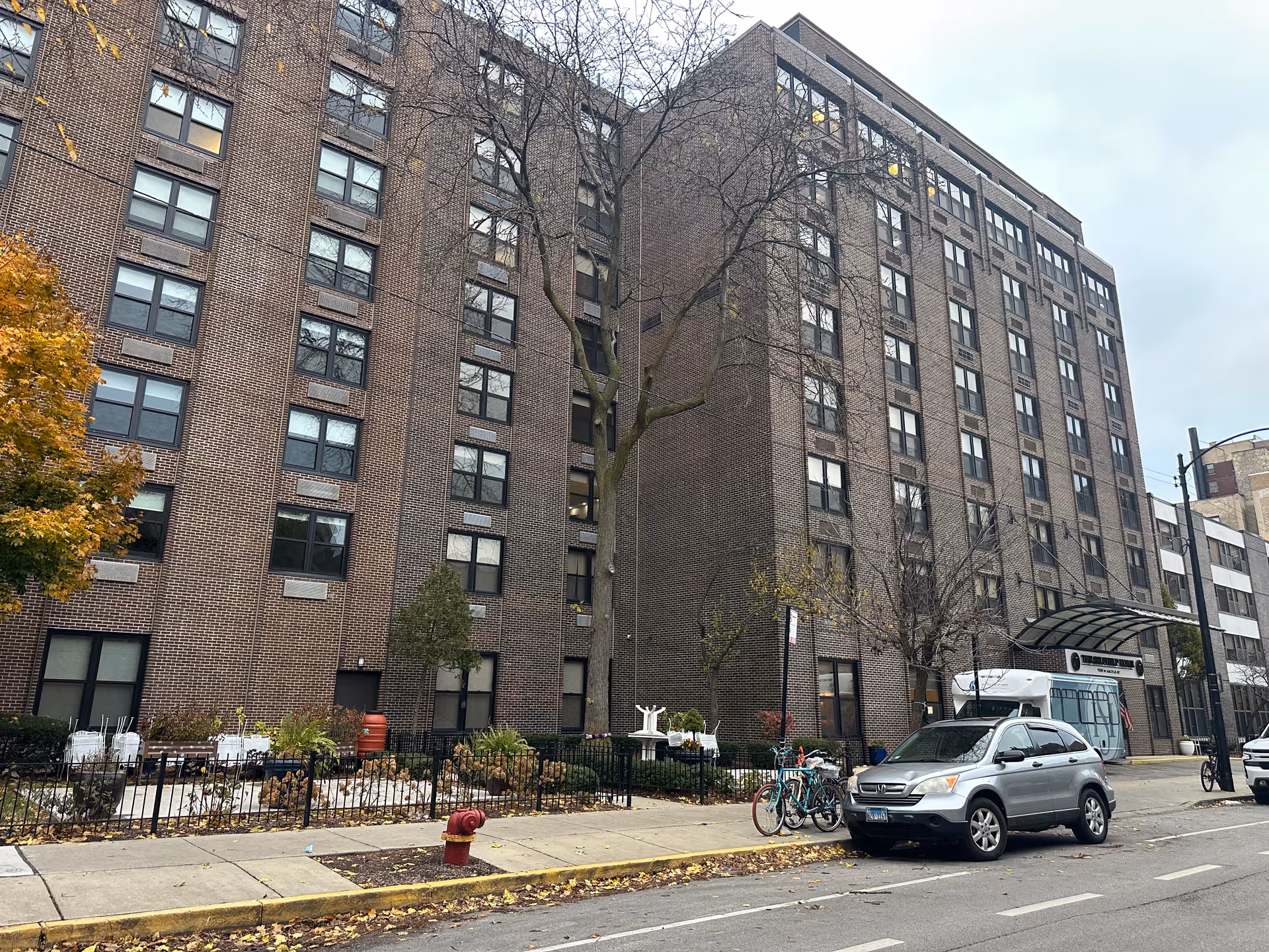 Exterior view of a multi-story brick building with numerous windows, a tree without leaves in front, a small fenced garden area, a silver SUV parked on the street, and a covered entrance with a sign above it. The sky is overcast.