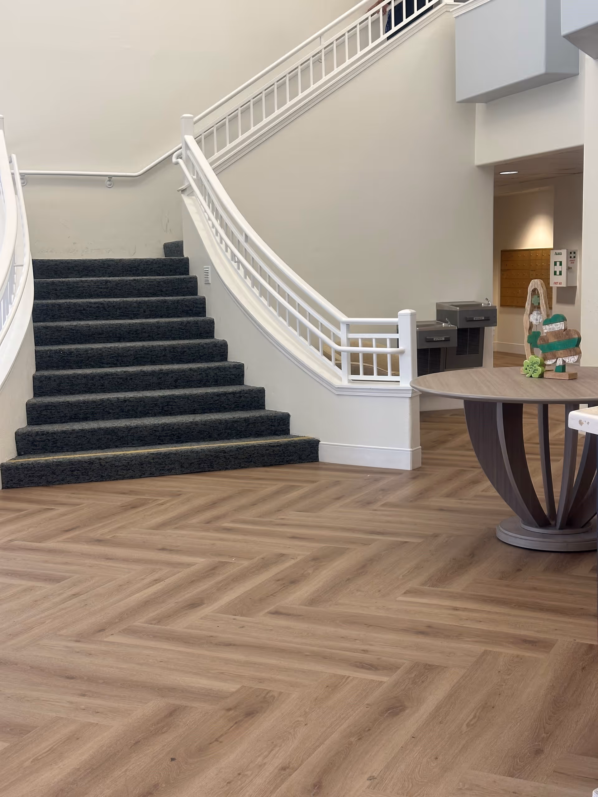 Interior view of a senior living facility showing a carpeted staircase with white railings, a wooden herringbone patterned floor, two water fountains mounted on the wall, and a round wooden table with decorative items on it.