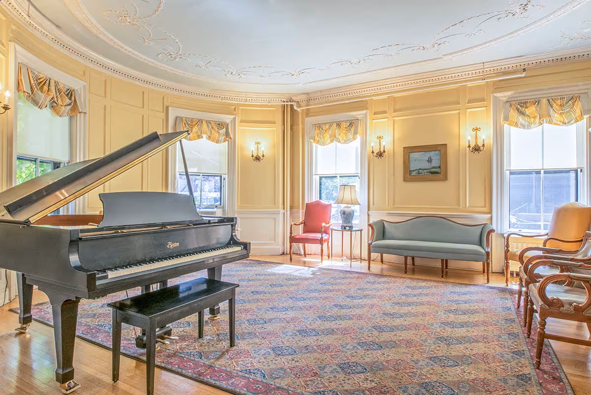 A spacious, elegant living room with a grand piano and bench on a large patterned rug. The room features yellow paneled walls, three large windows with striped valances, a red armchair, a blue upholstered sofa, wooden chairs, a side table with a lamp, and a framed painting on the wall. The ceiling has decorative molding.