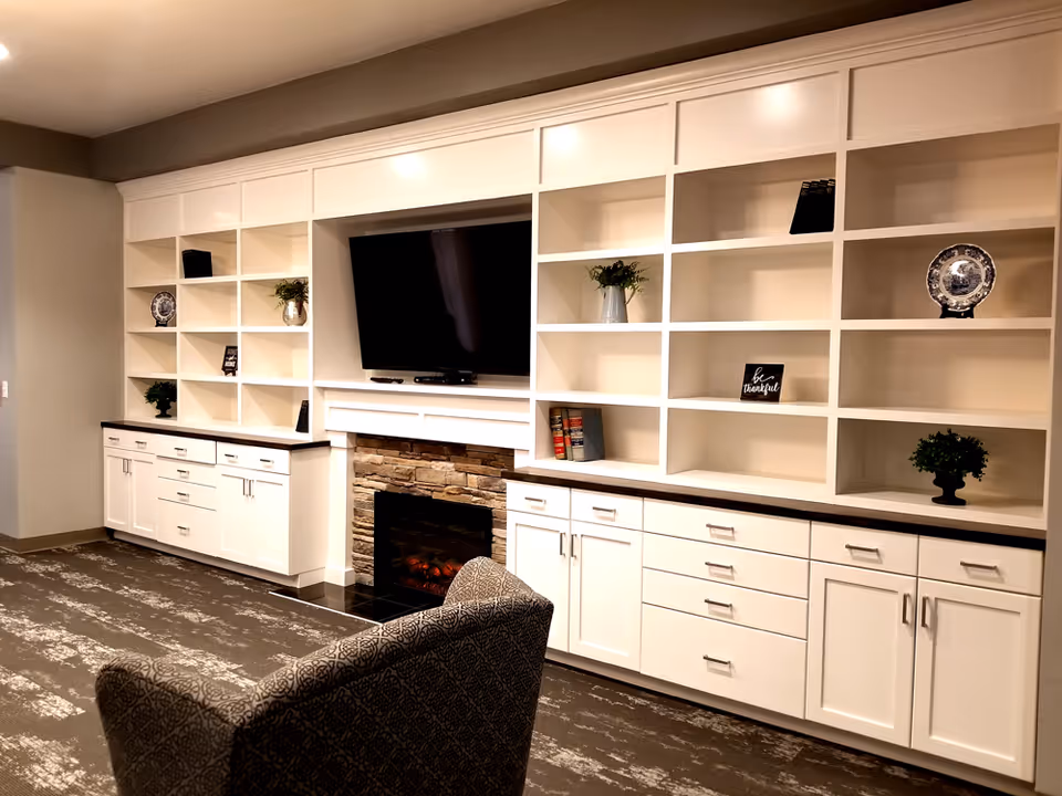 A cozy living room area with built-in white shelves and cabinets surrounding a flat-screen TV mounted above a stone fireplace. The shelves hold decorative items including plants, books, plates, and small signs. A patterned armchair is partially visible in the foreground.