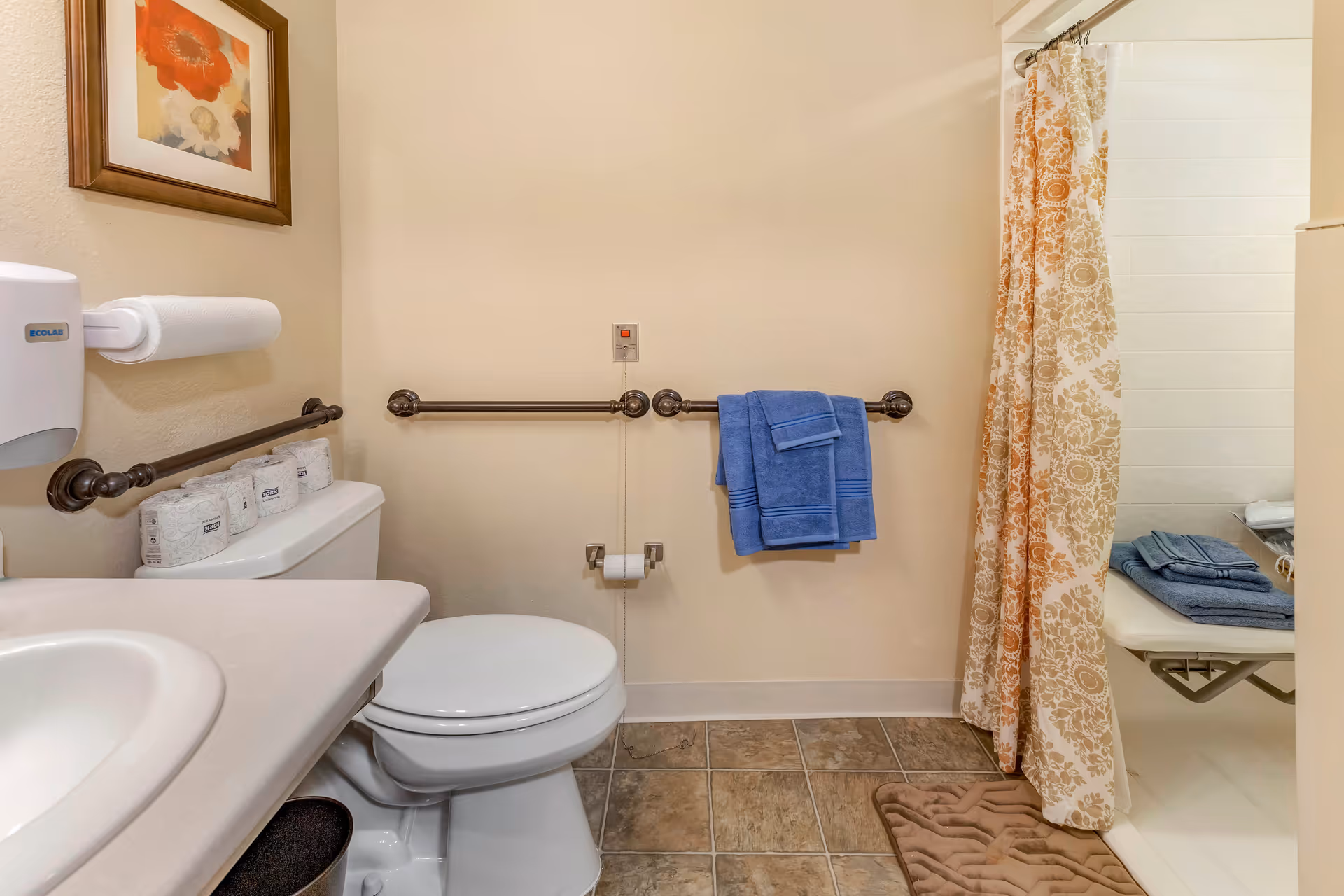 A bathroom with a toilet, sink, and shower. The toilet has three rolls of toilet paper on top. There are metal grab bars on the walls around the toilet, and a blue towel hanging on one of the bars. The shower has a patterned curtain in shades of orange and beige, and inside the shower, there is a fold-down seat with folded blue towels on it. The floor is tiled and there is a brown bath mat in front of the shower. A framed floral picture hangs on the wall above the toilet paper dispenser.