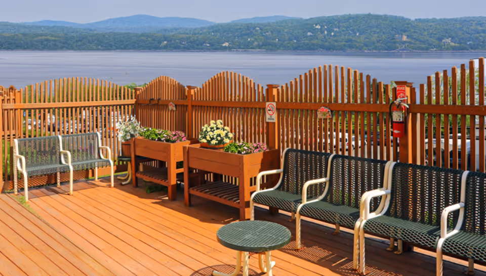 Outdoor wooden deck area with green metal benches and planters filled with flowers, overlooking a large body of water and distant tree-covered hills under a clear sky.