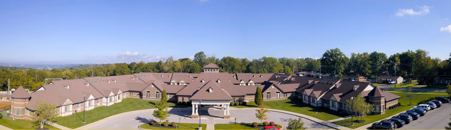 Aerial view of Forest Hills Healthcare Center, a large single-story building with a brown roof surrounded by green lawns, trees, and a parking lot with several cars. The sky is clear and blue.