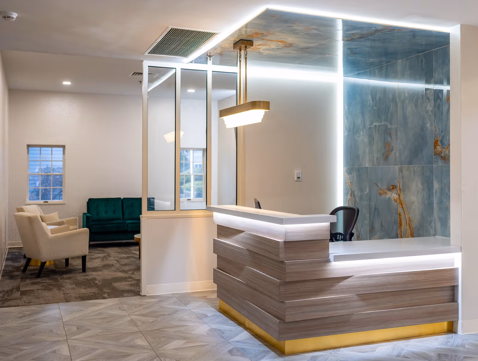 Modern reception area with a wooden front desk featuring built-in lighting, a decorative blue and gold wall behind the desk, and a hanging light fixture above. To the left, there is a seating area with beige armchairs and a green sofa near windows.