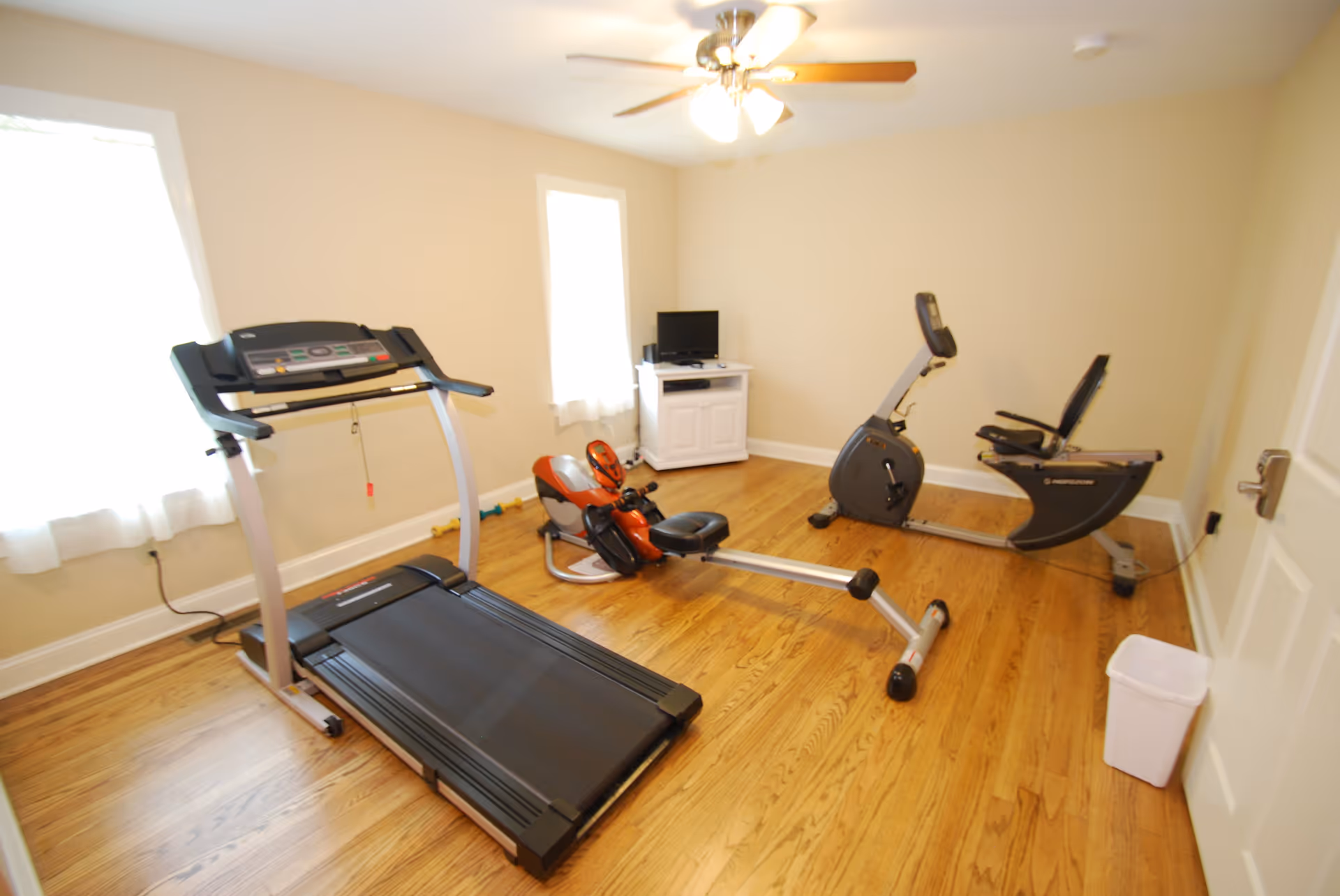 A small exercise room with wooden floors and beige walls featuring a treadmill, a rowing machine, and a recumbent exercise bike. There is a white cabinet with a small TV on top against the far wall, two windows with white curtains, a ceiling fan with lights, and a white trash bin near the door.