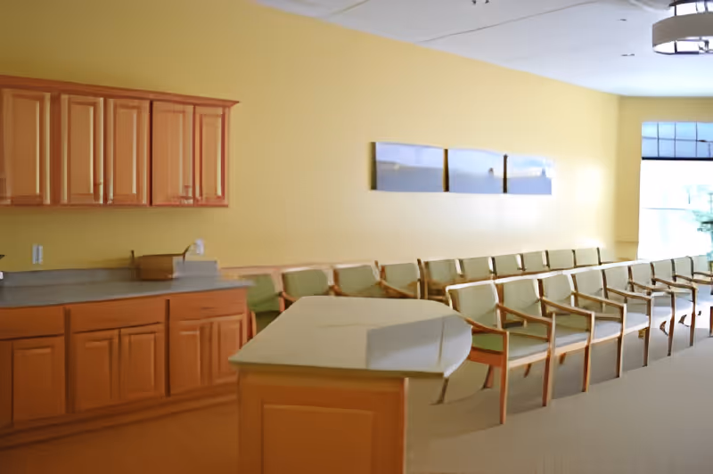 A waiting area with a row of green cushioned chairs with wooden frames lined up against a yellow wall. There is a countertop with wooden cabinets above and below on the left side of the image. Three landscape paintings are hung on the wall above the chairs. A large window is visible on the right side, allowing natural light into the room.